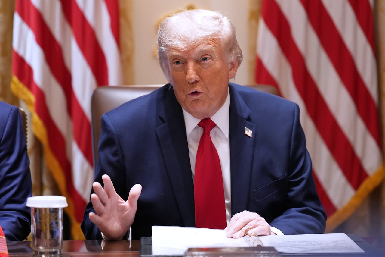 Image 4: An older blonde haired man gestures with his hand. He is wearing a suit and tie and seated in front of an American flag.